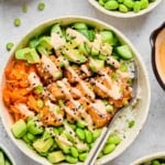 Salmon rice bowl with cubed salmon, white rice, edamame, diced avocado, shredded carrots, and diced cucumber with a fork in the bowl. Everything is drizzled with sriracha mayo and garnished with sesame seeds and green onions. Around the bowl is another rice bowl, a bowl of diced cucumber, a bowl of shredded carrots, a bowl of edamame, and a dish of sriracha mayo.