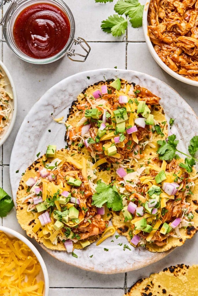 Overhead shot of three bbq chicken tacos on a plate. The tacos are filled with coleslaw, avocado, shredded cheese, red onion, and cilantro. Around the plate is a bowl of shredded bbq chicken, corn tortillas, a bowl of shredded cheese, and a jar of bbq sauce.