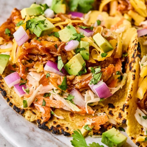 Bbq chicken tacos in a corn tortilla with coleslaw, shredded bbq chicken, avocado, shredded cheese, red onion, and cilantro on a plate.