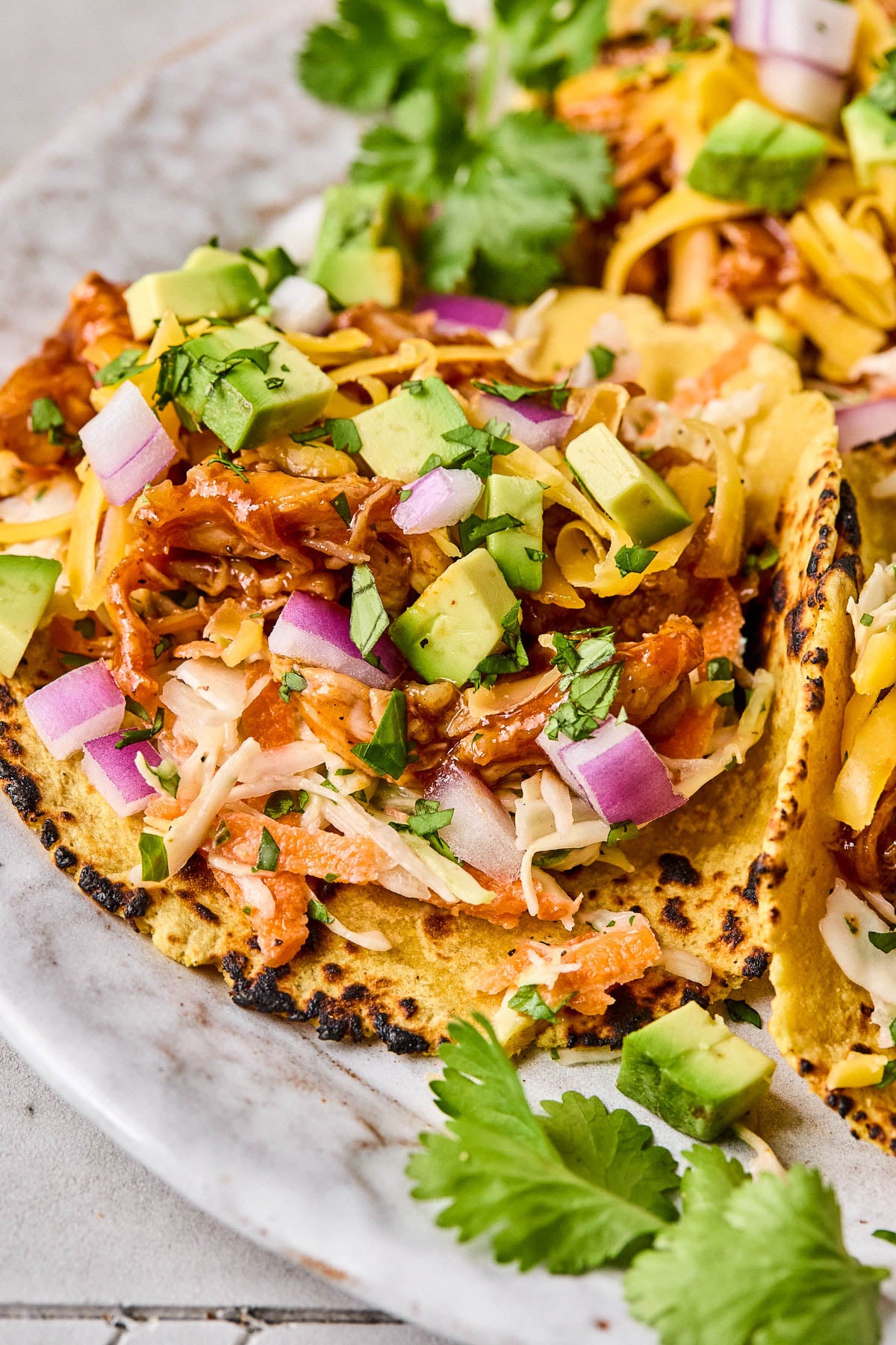 Bbq chicken tacos in a corn tortilla with coleslaw, shredded bbq chicken, avocado, shredded cheese, red onion, and cilantro on a plate.