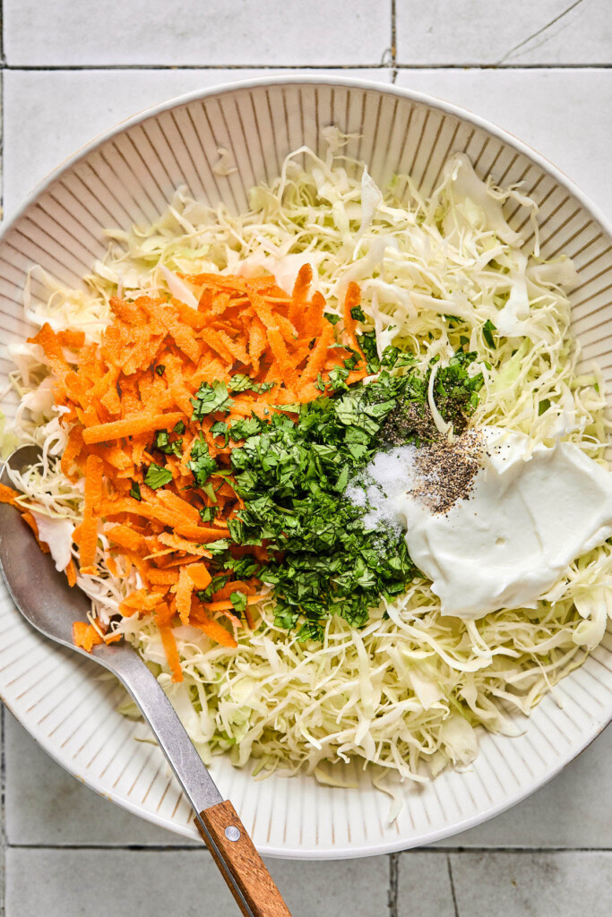 Large white bowl with shredded cabbage, shredded carrots, greek yogurt, cilantro, salt, and pepper before it is all mixed together. A large spoon is also in the bowl.