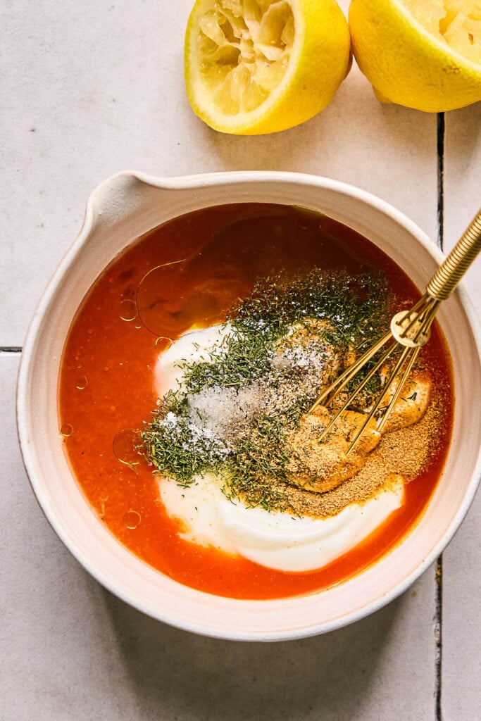 A bowl with hot sauce, yogurt, spices, salt, and pepper in it before it is mixed together. A small whisk is in the bowl and a juiced lemon is next to the bowl.