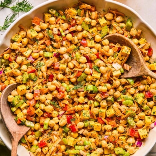 Buffalo chickpea salad with veggies, cheese, and herbs in a large bowl with two wooden spoons in it. Next to the bowl is fresh dill, a lemon that has been cut in half and juiced, a jar of dressing, and a green kitchen linen.