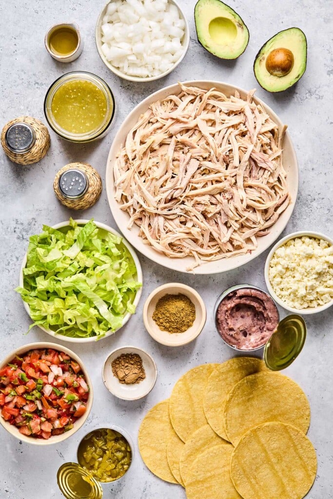 Grey concrete counter with an avocado cut in half, a large bowl of shredded chicken, a bowl of crumbled queso fresco, an opened can of refried black beans, corn tortillas, two small bowls of spices, an opened can of diced green chiles, a bowl of pico de gallo, a bowl of shredded lettuce, a salt and pepper shaker, a bowl of green salsa, and a small container of olive oil.