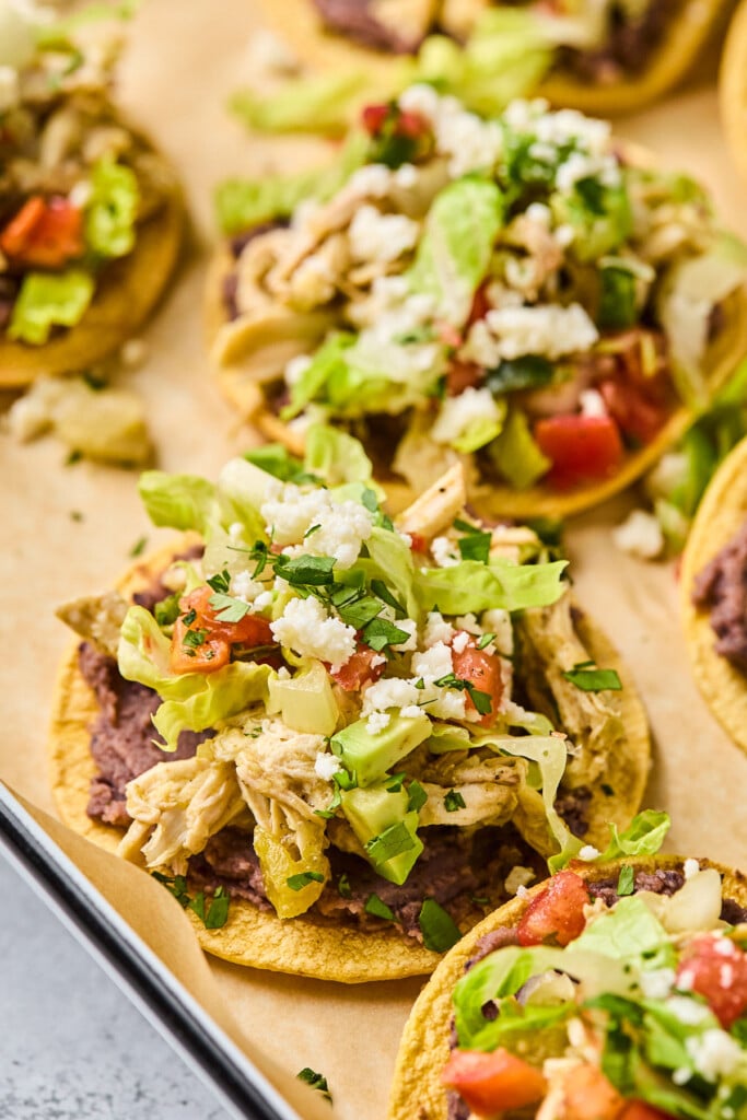 Shredded chicken tostadas with refried beans, shredded lettuce, pico de gallo, crumbled queso fresco, and cilantro. They are spread across a baking sheet.
