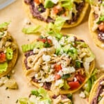 Chicken tostadas with refried beans, seasoned shredded chicken, lettuce, pico de gallo, queso fresco, and cilantro on them. They are on a baking sheet lined with parchment paper.