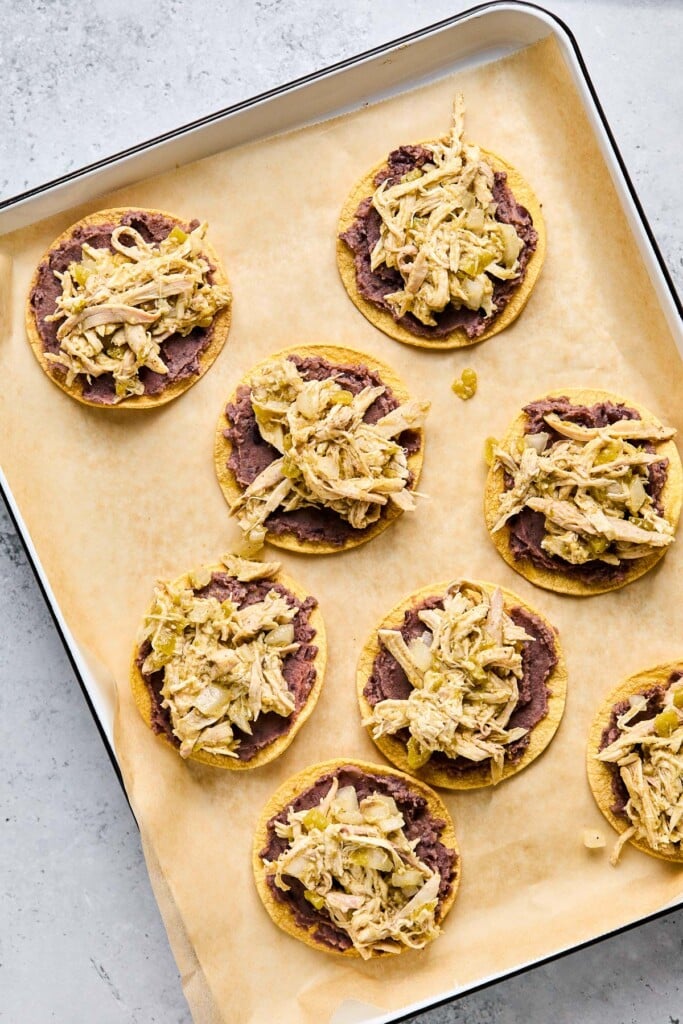 Overhead shot of a baking sheet lined with parchment paper with crispy corn tortilla shells with refried beans and seasoned shredded chicken on them.