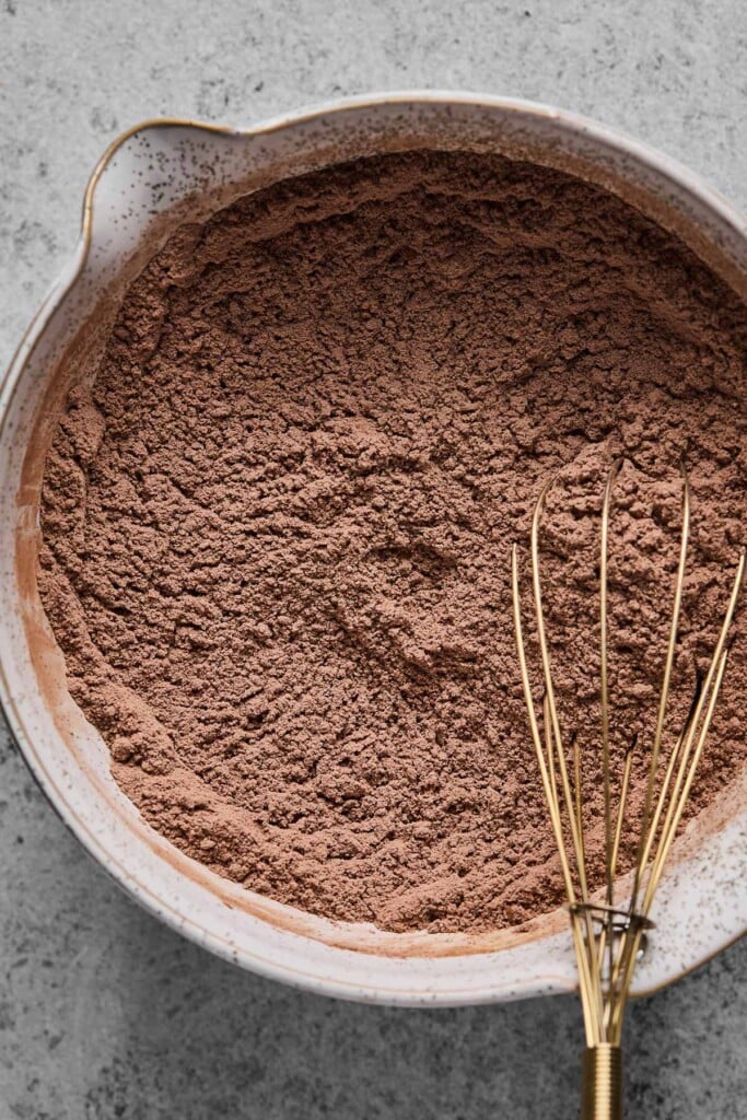 An overhead shot of a mixing bowl with flour, chocolate protein powder, cocoa powder, and spices mixed together. A whisk is also in the bowl.