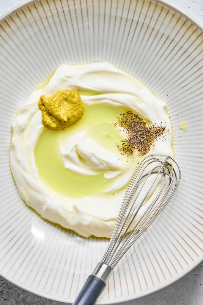 A large bowl with greek yogurt, dijon mustard, pickle juice, and spices in it before it is mixed together. A whisk is also in the bowl.