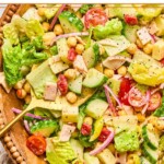 Chopped salad with lettuce, tomatoes, cucumber, turkey, cheese, chickpeas, red onion, and banana peppers in a large bowl. It is garnished with pepper and there is a large gold spoon in the bowl as well.
