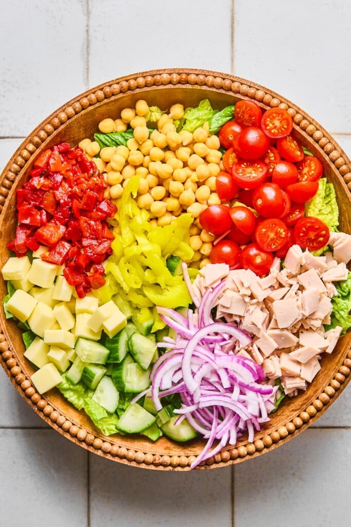 Large brown bowl with a bed of chopped lettuce with halved cherry tomatoes, chopped deli turkey, sliced red onions, diced cucumbers, cubed cheese, chopped roasted red peppers, chickpeas, and banana peppers in separate piles on top of it.