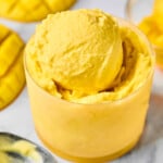 Scoops of mango Ninja Creami frozen yogurt in a small glass cup. Around the cup is an ice cream scoop and fresh mango that has been cut from the seed.