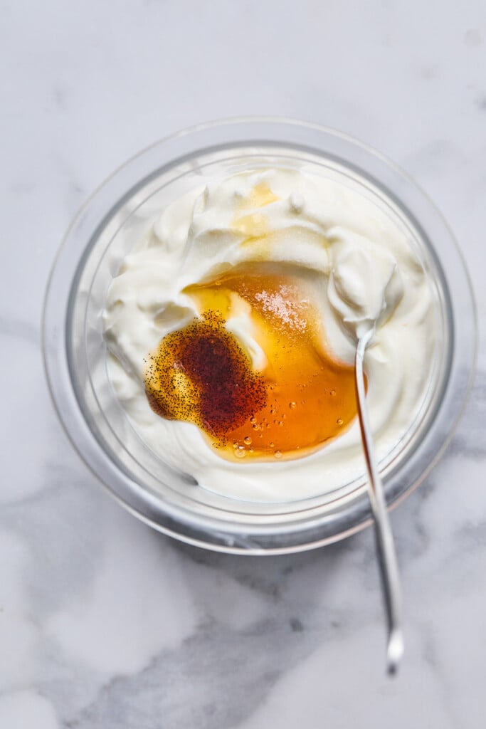 Pint container with greek yogurt, honey, vanilla bean paste, and salt before it is mixed together. A spoon is also in the container.