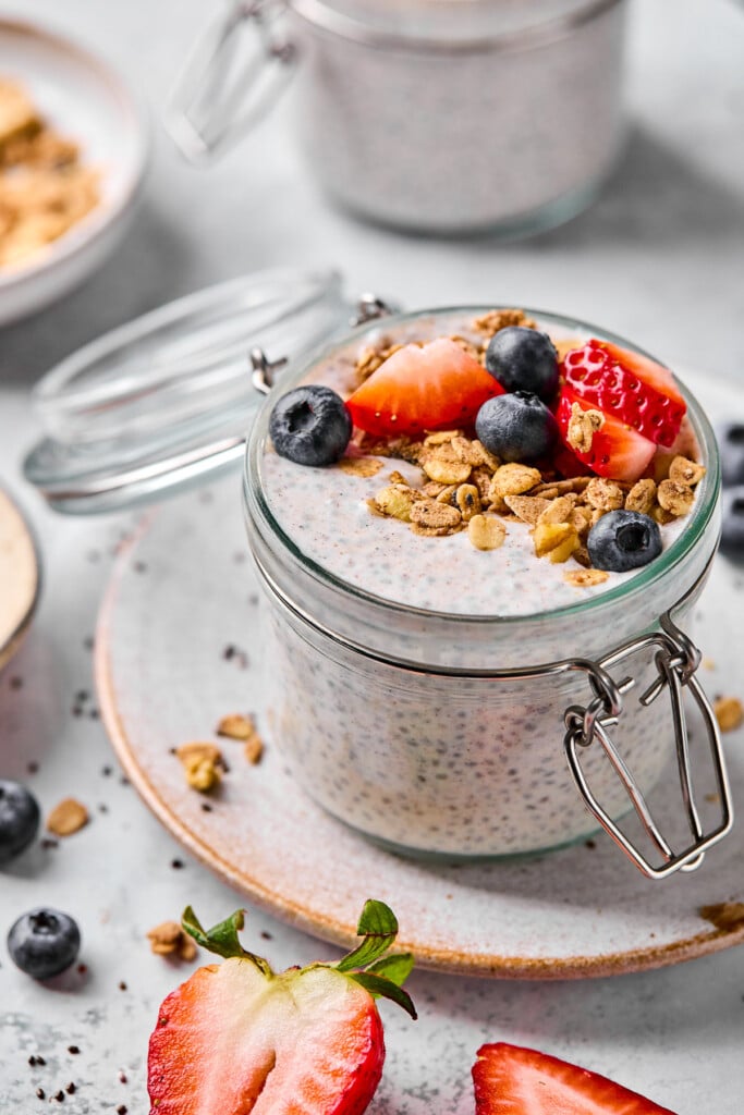A jar of high protein chia pudding topped with strawberries, blueberries, and granola. The jar is sitting on a plate. Around the plate are some berries, a bowl of granola, and another jar of chia pudding.