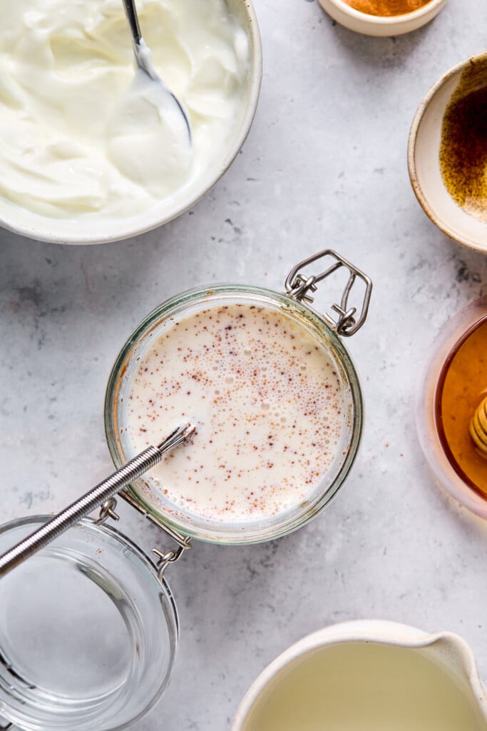 Overhead shot of a jar with a mixture of greek yogurt, milk, honey, and cinnamon in it. A whisk is also in the jar. Around the jar is a bowl of greek yogurt, a bowl of vanilla bean paste, a bowl of honey, and a cup of milk.