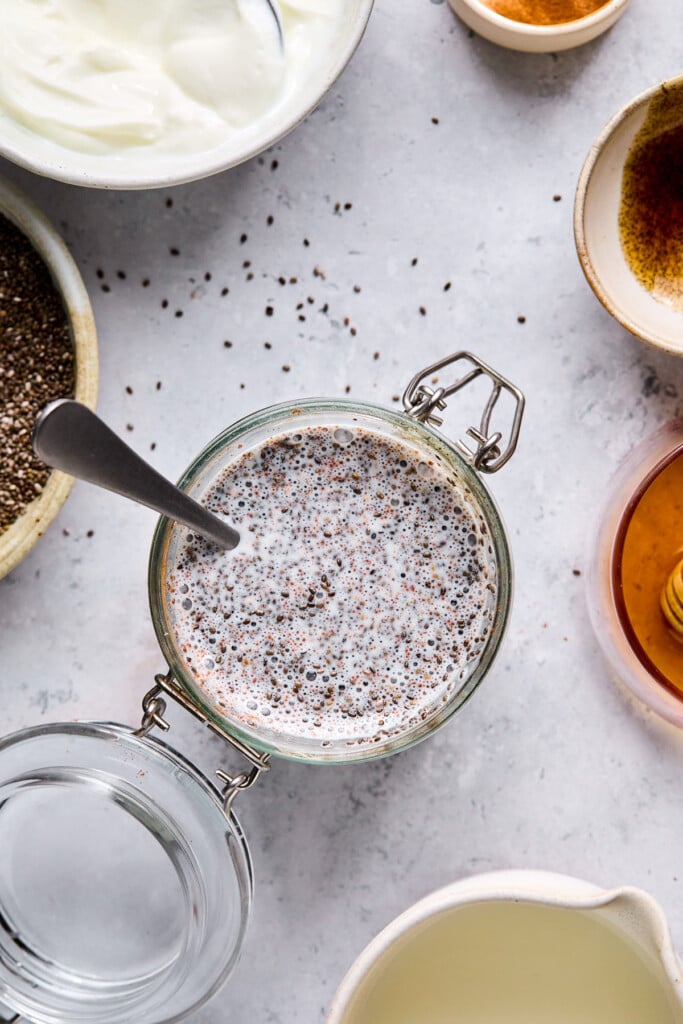 Overhead shot of a jar with a mixture of greek yogurt and milk mixture with chia seeds before it gels. A spoon is also in the jar. Around the jar is a bowl of greek yogurt, a bowl of vanilla bean paste, a bowl of honey, a cup of milk, and a bowl of chia seeds.