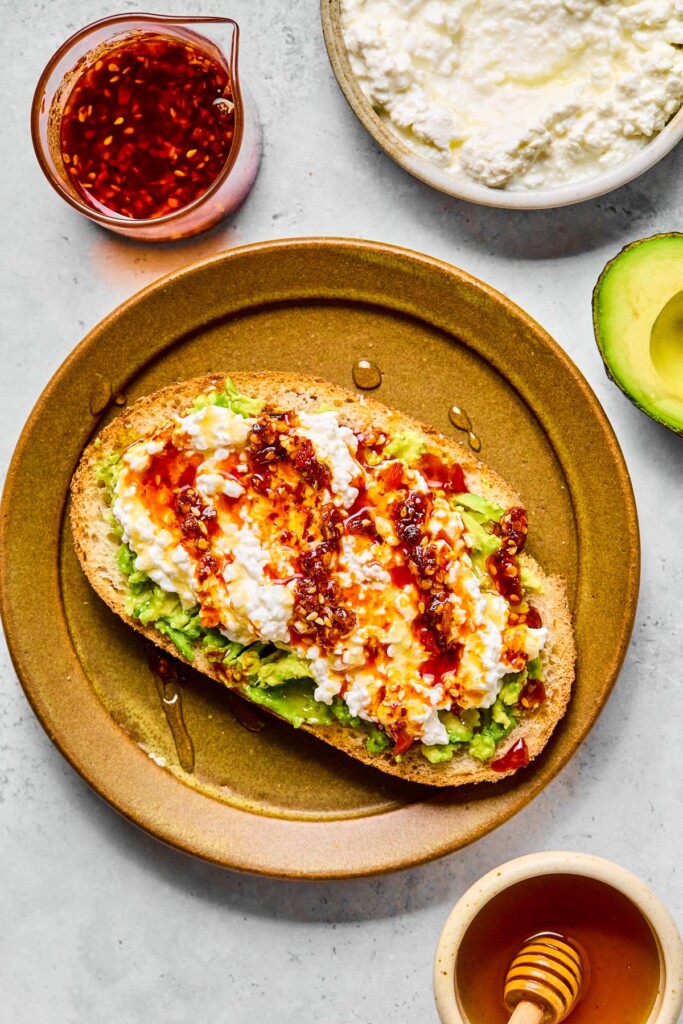 Avocado toast with cottage cheese, chili oil, and honey on a brown plate. Around the plate is a bowl of cottage cheese, half an avocado, a small bowl of honey with a honey dipper in it, and a cup of chili oil.