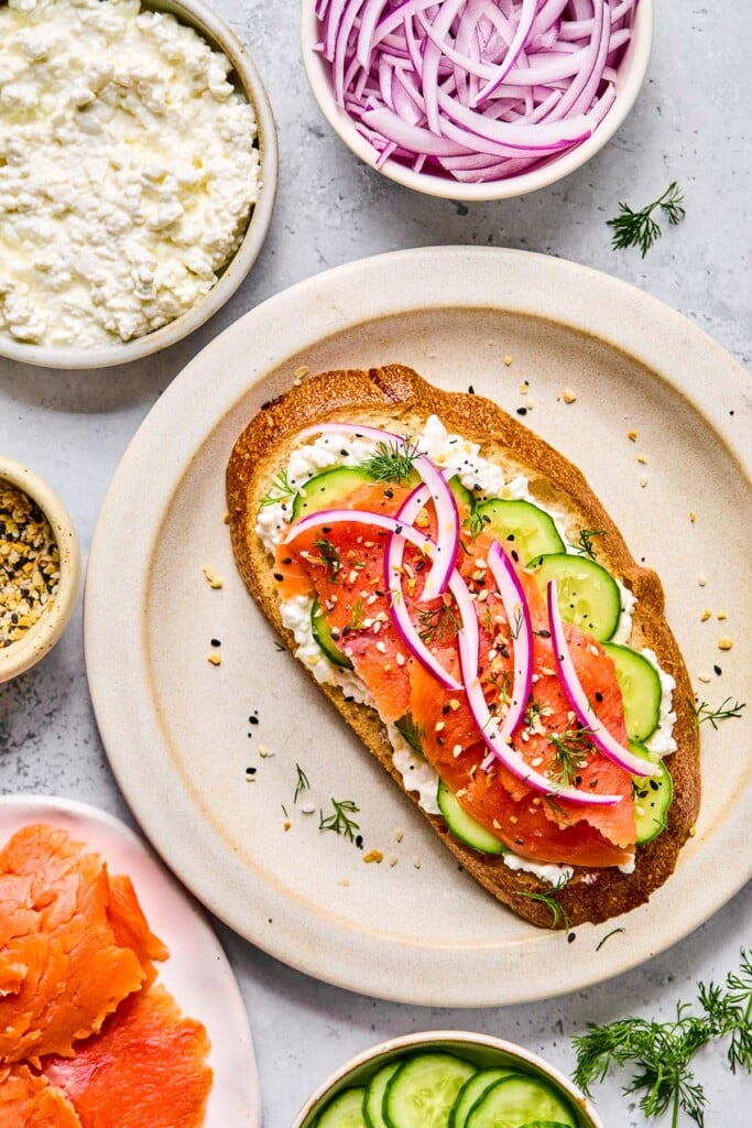 Smoked salmon cottage cheese toast with sliced cucumbers, sliced red onion, everything but the bagel seasoning, and fresh dill on a plate. Around the plate is a bowl of sliced red onion, fresh dill, a bowl of sliced cucumber, a plate of smoked salmon, a bowl of everything but the bagel seasoning, and a bowl of cottage cheese.