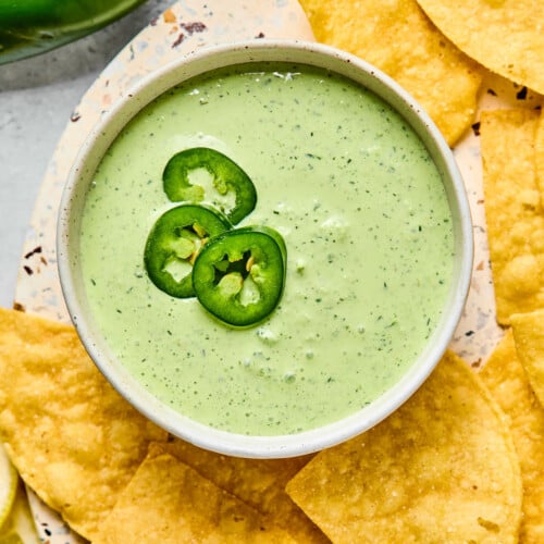 A bowl of jalapeno ranch garnished with sliced jalapenos. The bowl is on a plate that is filled with tortilla chips. Next to the plate are some whole jalapenos.