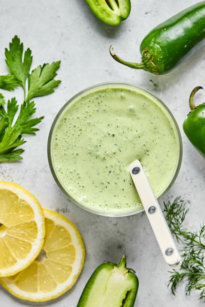 A glass cup filled with jalapeno ranch dip, with a spoon in it. Around the cup are lemon slices, a sprig of fresh parsley, jalapenos, and a sprig of fresh dill.