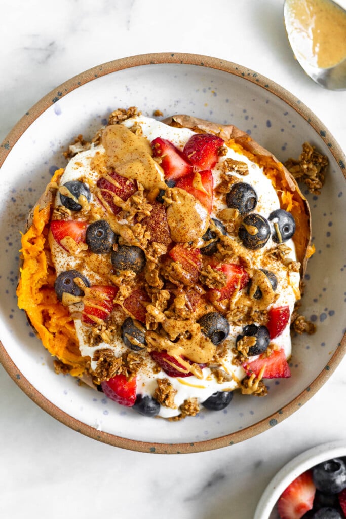 Greek yogurt stuffed a cooked sweet potato topped with berries, granola, nut butter, and cinnamon in a bowl. Next to the bowl is a bowl of berries and a spoon with peanut butter on it.