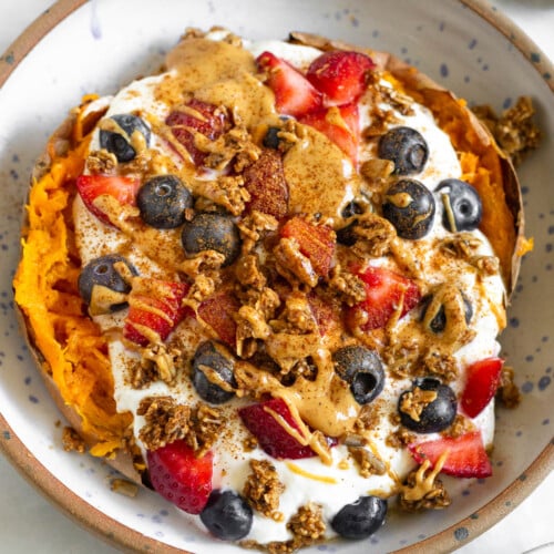 Sweet potato breakfast bowl with greek yogurt, berries, nut butter, granola, and topped with cinnamon. Next to the bowl is a bowl of berries and a spoon with peanut butter on it.