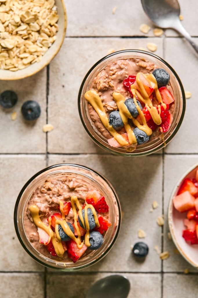 An overhead shot of two jars of chocolate overnight oats topped with blueberries, diced strawberries, and peanut butter. Next to the jars is a bowl of diced strawberries, some blueberries, two spoons, and a bowl of rolled oats.
