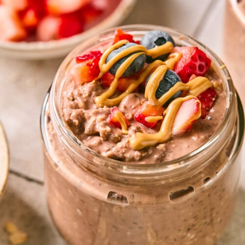 Jar of chocolate overnight oats topped with diced strawberries, blueberries, and a drizzle of peanut butter. Around the jar are some rolled oats, a blueberry, a bowl of diced strawberries, and another jar of overnight oats.