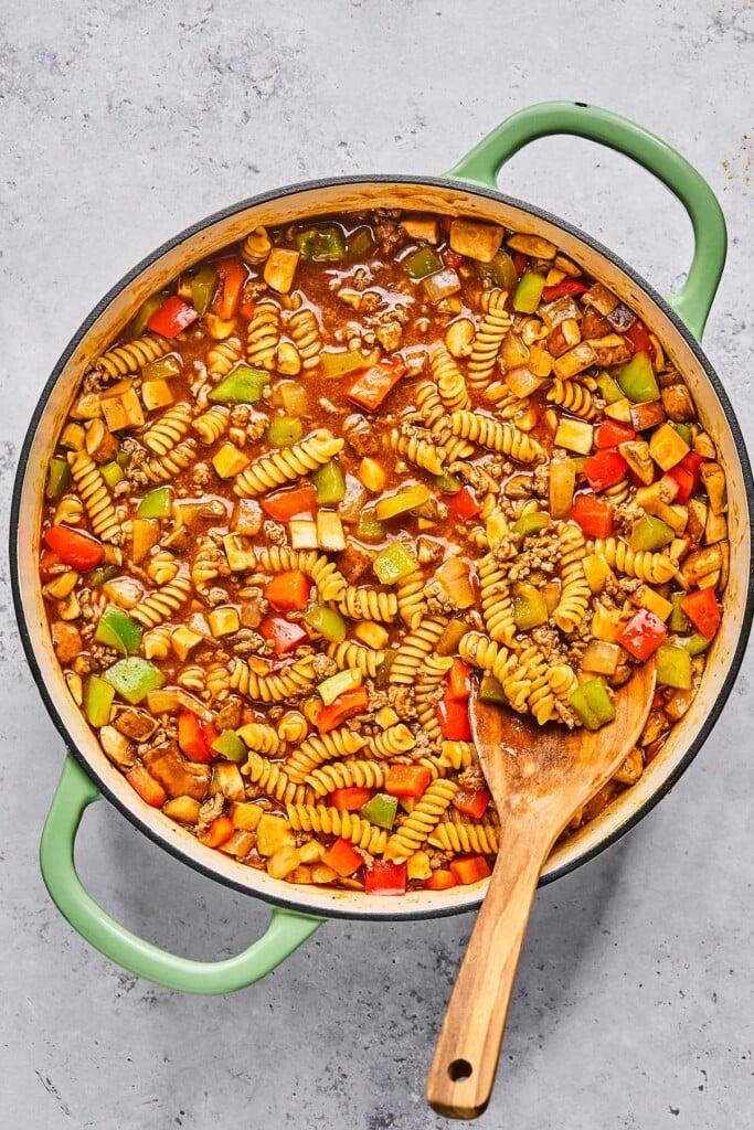 Large enameled pan filled with meat, veggies, and pasta in a red sauce. A wooden spoon is in the pot.