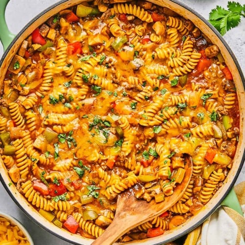 Overhead shot of enchilada pasta in a large pot topped with melted cheese and cilantro. A wooden spoon is also in the pot. Around the pot is a a plate of lime wedges, some sprigs of fresh cilantro, a tan kitchen towel, and a bowl of shredded cheddar cheese.
