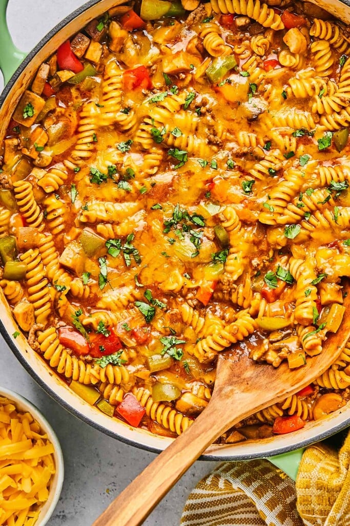 One pot enchilada pasta with ground beef, veggies, and melted cheese in a large enameled pan. It is garnished with cilantro and there is wooden spoon in the pan. Around the pan is a tan kitchen towel and a bowl of shredded cheese.