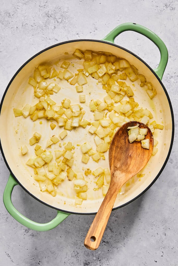 Large enameled pan filled with sautéed onions and garlic. There is a wooden spoon in the pan as well.