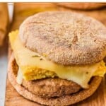 Freezer breakfast sandwich with sausage, egg, and cheese on an English muffin on a wooden cutting board. There are some sandwiches in the background.