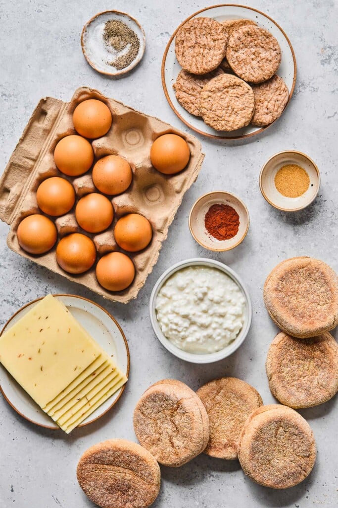 Grey concrete counter with a plate of frozen chicken sausage patties, a small bowl of garlic powder, a small bowl of paprika, a bowl of cottage cheese, 6 English muffins, a plate with slices of cheese on it, a large egg carton with 10 eggs in it, and a small bowl of salt and pepper on it.