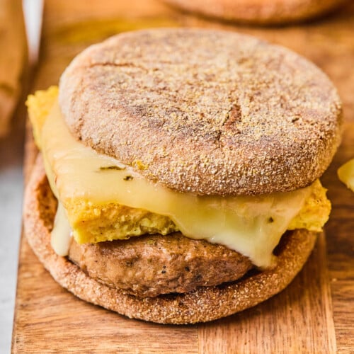 Freezer breakfast sandwich with sausage, egg, and cheese on an English muffin on a wooden cutting board. There are some sandwiches in the background.