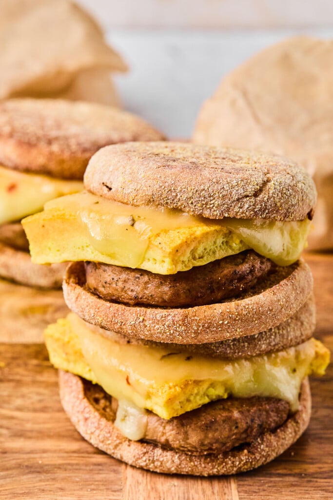 Two meal prep breakfast sandwiches stacked on top of each other on a wooden cutting board. Behind them are more breakfast sandwiches.