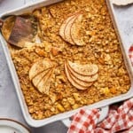 A plan of apple cinnamon baked oatmeal with a piece cut out of it. A spatula is in the pan where the pieces is gone. Around the pan is a a plate of chopped walnuts, a red and white checkered towel, a plate with a fork and a piece of the baked oatmeal on it, and some red apples.