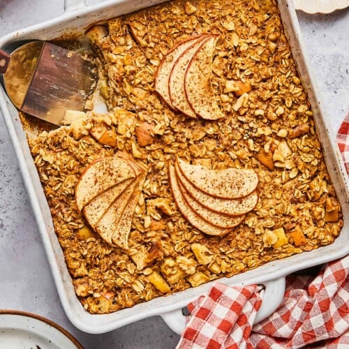 A plan of apple cinnamon baked oatmeal with a piece cut out of it. A spatula is in the pan where the pieces is gone. Around the pan is a a plate of chopped walnuts, a red and white checkered towel, a plate with a fork and a piece of the baked oatmeal on it, and some red apples.