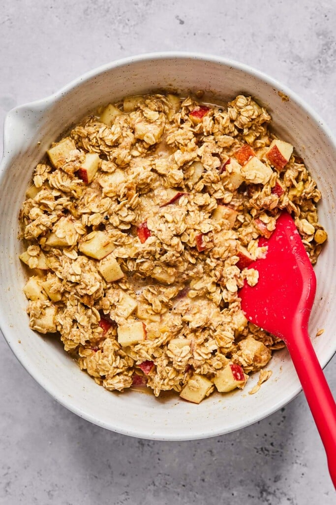 A mixing bowl filled with a batter of rolled oats with diced apples and walnuts. A red spatula is in the bowl as well.