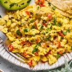 A plate of migas, sliced avocado, and tortillas. The migas are garnished with salsa and cilantro. Next to the plate is a green kitchen towel and another plate of migas.
