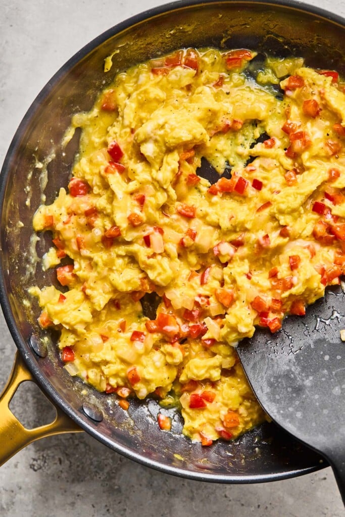 Eggs being scrambled with diced peppers and onions in a large skillet. A spatula is off to the side in the skillet.