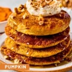 A stack of pumpkin protein pancakes topped with greek yogurt, pecan pieces, and maple syrup on a plate. Around the plate are some pecan pieces, cinnamon sticks, a bowl of pumpkin puree, and bowl of pecans.