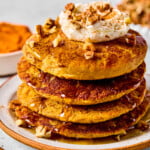 A stack of pumpkin protein pancakes topped with greek yogurt, pecan pieces, and maple syrup on a plate. Around the plate are some pecan pieces, cinnamon sticks, a bowl of pumpkin puree, and bowl of pecans.