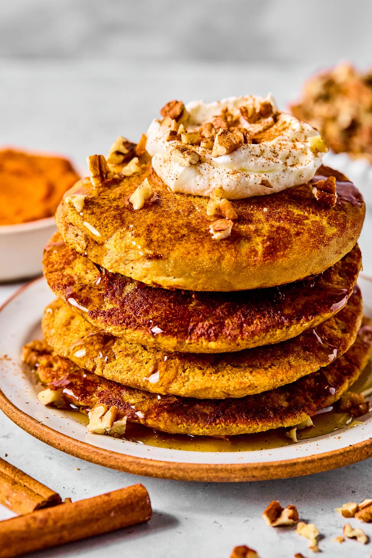 A stack of pumpkin protein pancakes topped with greek yogurt, pecan pieces, and maple syrup on a plate. Around the plate are some pecan pieces, cinnamon sticks, a bowl of pumpkin puree, and bowl of pecans.