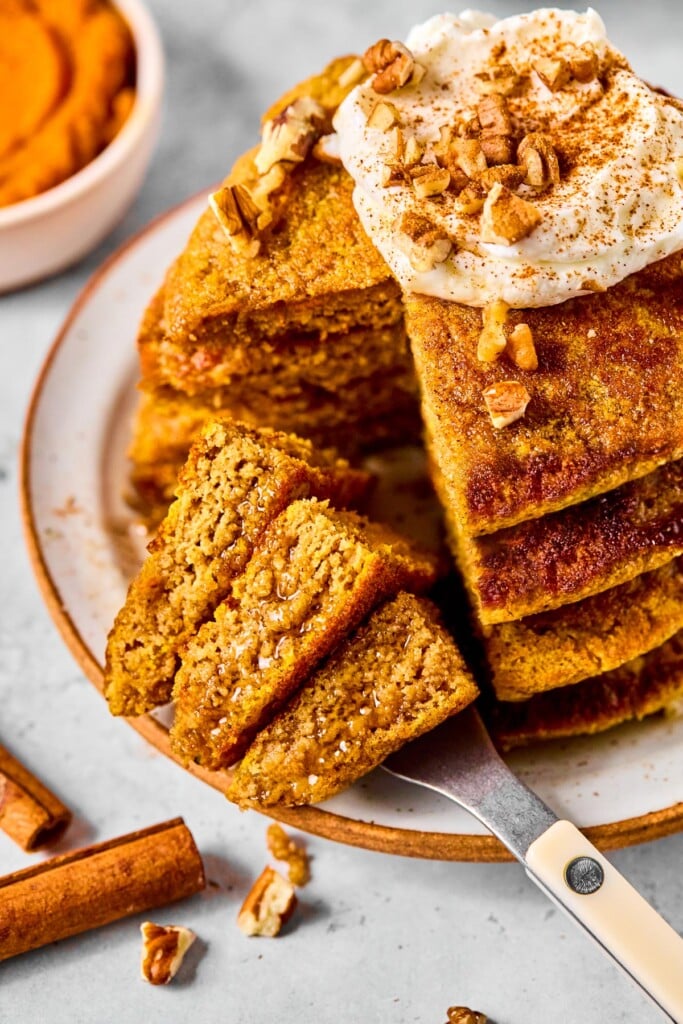 High protein pumpkin pancakes stacked on a plate with a section cut out of them on a fork. They are topped with yogurt, pecan pieces, maple syrup, and cinnamon. Around the plate are some cinnamon sticks, pecan pieces, and a bowl of pumpkin puree.