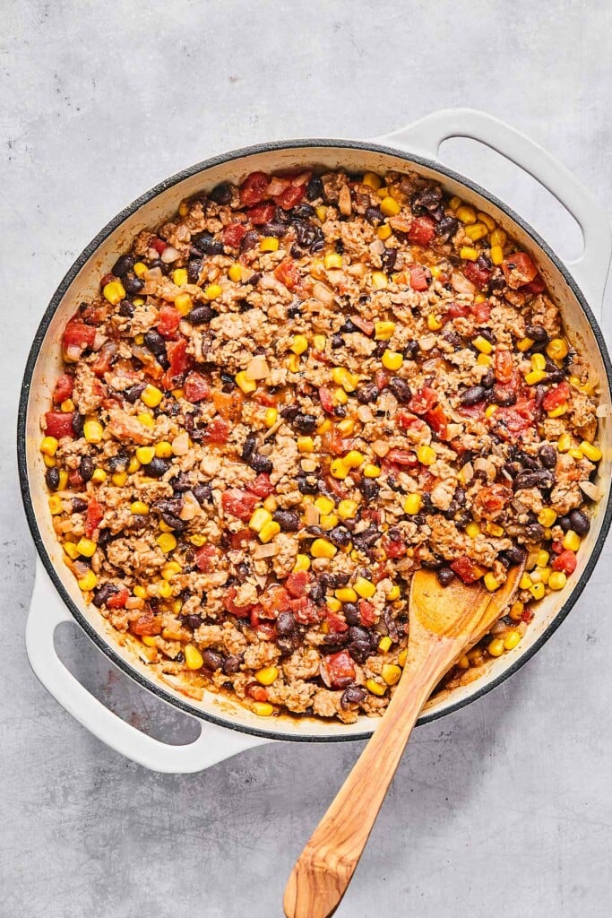 Large enameled white with a mixture of ground turkey, black beans, diced tomatoes, corn, and spices in it. There is a wooden spoon in the pan as well.