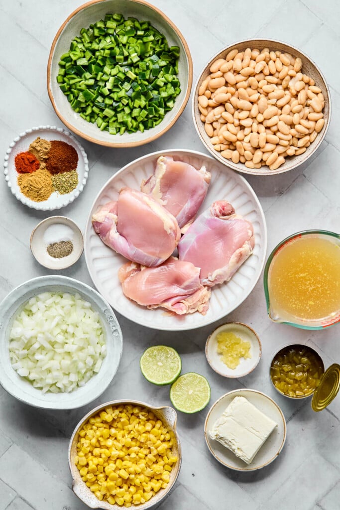 A bowl of white beans, a plate of four raw chicken thighs, a measuring cup of broth, a small dish of minced garlic, an open can of green chiles, a plate with half a brick of cream cheese on it, a lime that has been cut in half, a bowl of canned corn, a bowl of diced onion, a small bowl of salt and pepper, a small plate with 5 different spices on it, and a bowl of diced poblano pepper on a grey counter.