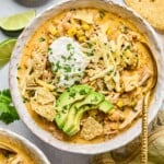 A bowl of healthy white chicken chili topped with greek yogurt, sliced avocado, shredded cheese, tortilla chips, and cilantro. There is a spoon in the bowl as well. Around the bowl are some tortilla chips, a tan kitchen towel, another bowl of spoon, some lime wedges, and a plate of chopped cilantro.