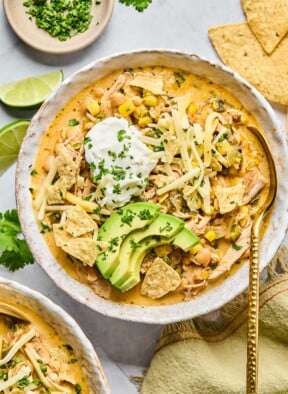 A bowl of healthy white chicken chili topped with greek yogurt, sliced avocado, shredded cheese, tortilla chips, and cilantro. There is a spoon in the bowl as well. Around the bowl are some tortilla chips, a tan kitchen towel, another bowl of spoon, some lime wedges, and a plate of chopped cilantro.