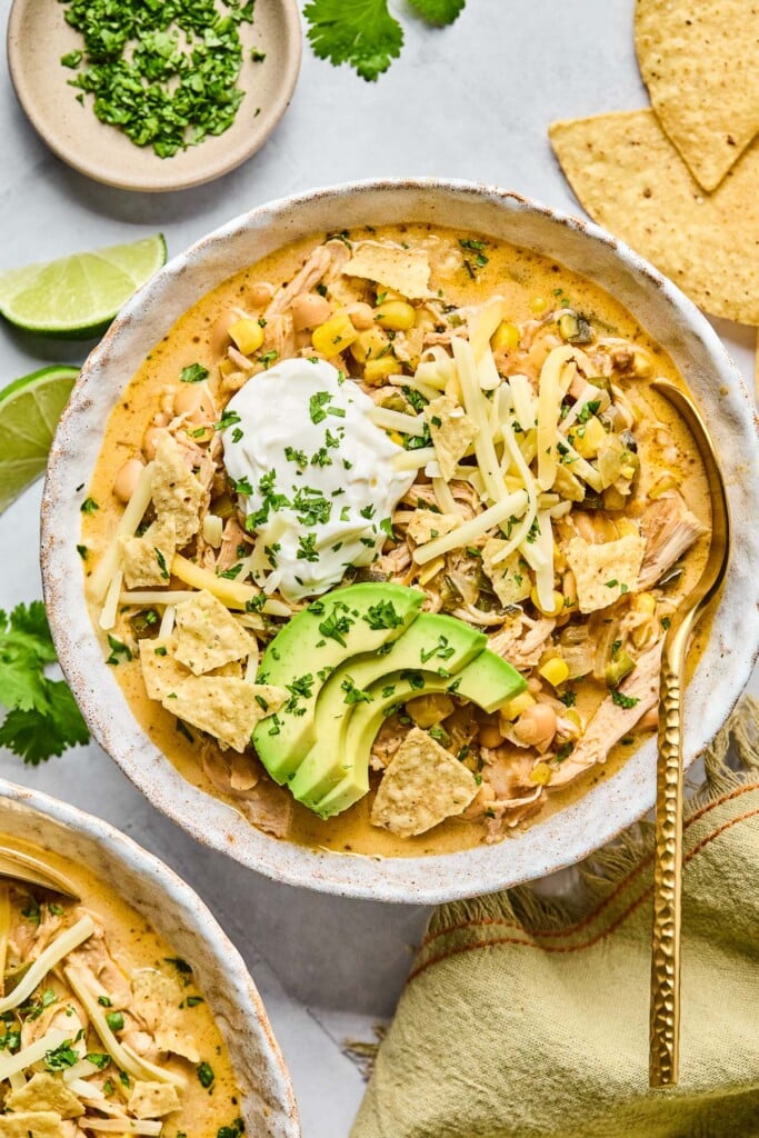 A bowl of healthy white chicken chili topped with greek yogurt, sliced avocado, shredded cheese, tortilla chips, and cilantro. There is a spoon in the bowl as well. Around the bowl are some tortilla chips, a tan kitchen towel, another bowl of spoon, some lime wedges, and a plate of chopped cilantro.